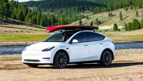 White Tesla Model Y parked at a lake with a red surfboard on top. Tesla has increased the price of the Model Y and it could be because of new EV tax credits