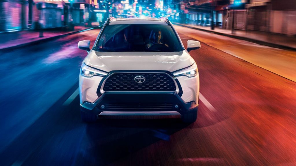 White 2022 Toyota Corolla Cross driving on a city street at night, the compact crossover SUV crushes the 2022 Nissan Rogue Sport, according to KBB.