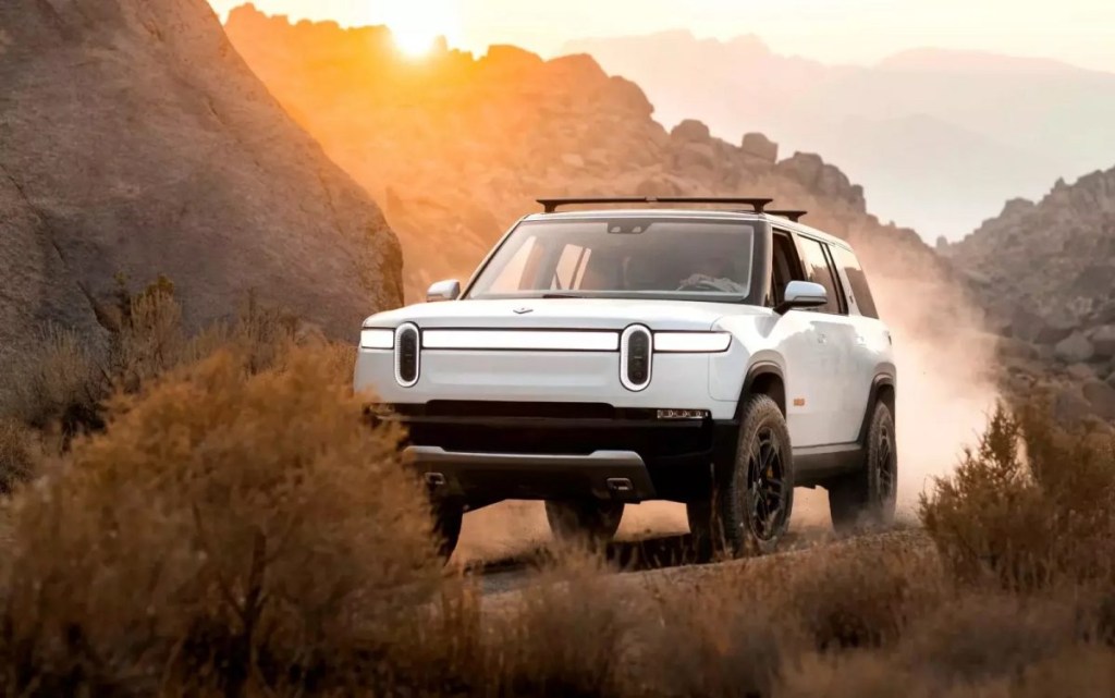White 2022 Rivian R1S, with price info, driving on a dirt road by a large rock
