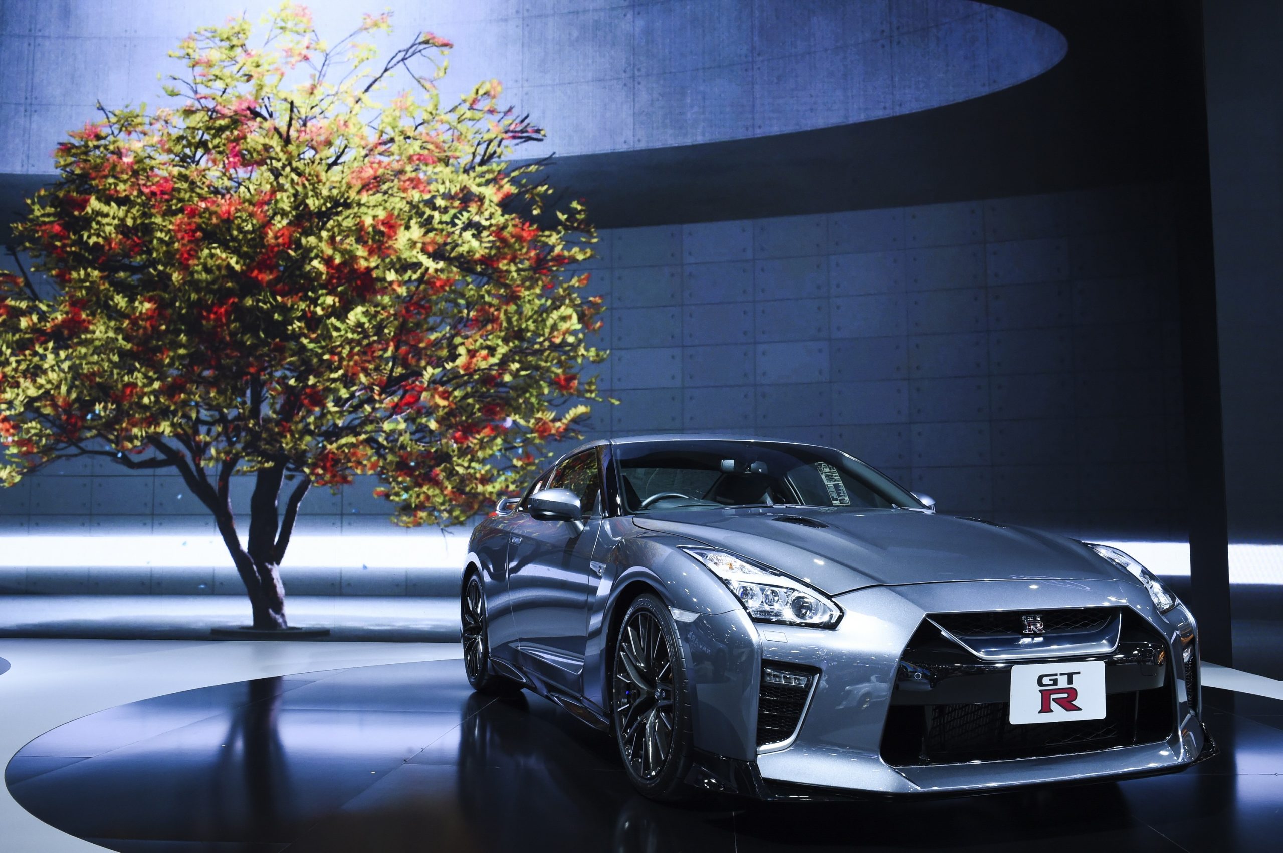 A silver Nissan GT-R shot from the front 3/4 in front of a tree in a display booth