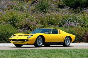 A 1970 Lamborghini Miura P400S supercar parked on a road in front of a wildflower-dotted hill