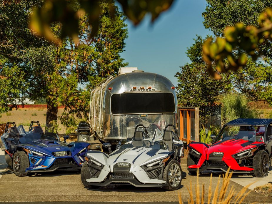 A blue 2022 Polaris Slingshot R next to a white 2022 SL next to an accessorized red 2022 S in front of an Airstream trailer