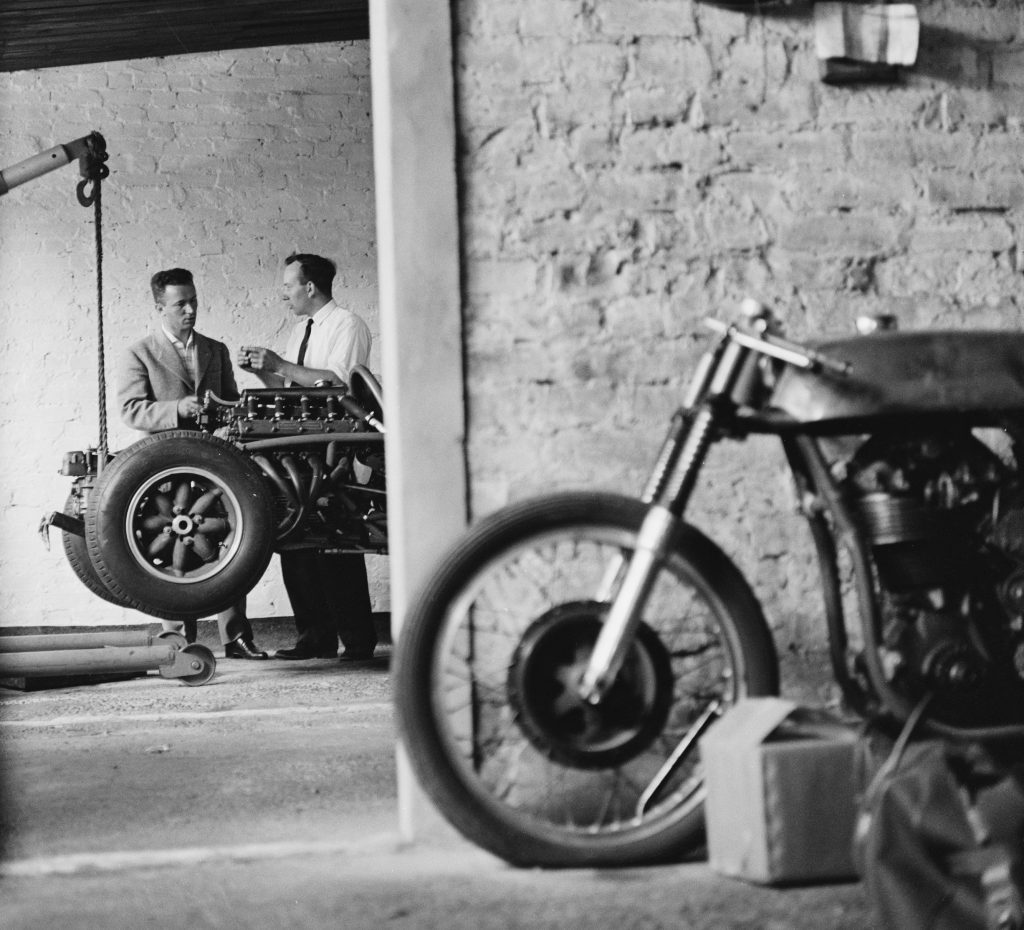 Arturo Magni and John Surtees discuss Surtees' Formula Two Cooper next to a motorcycle in a garage