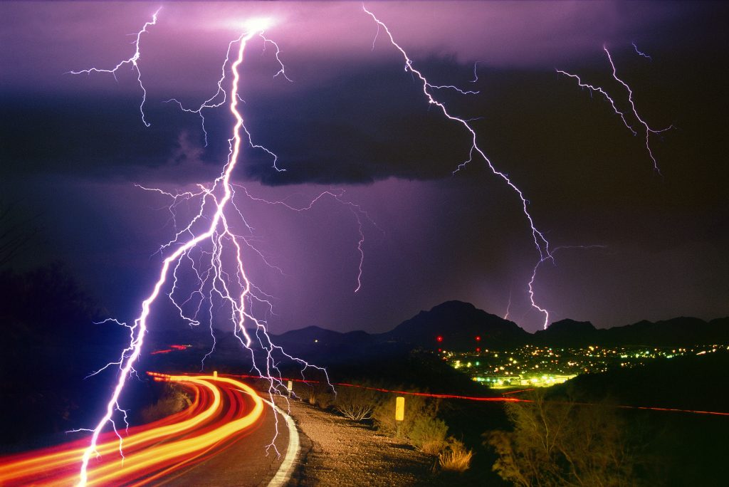 Sonoran Desert rainstorm, a summer monsoon thunderstorm with cloud-to-ground lightning strikes and light trails from cars on the roadway.
