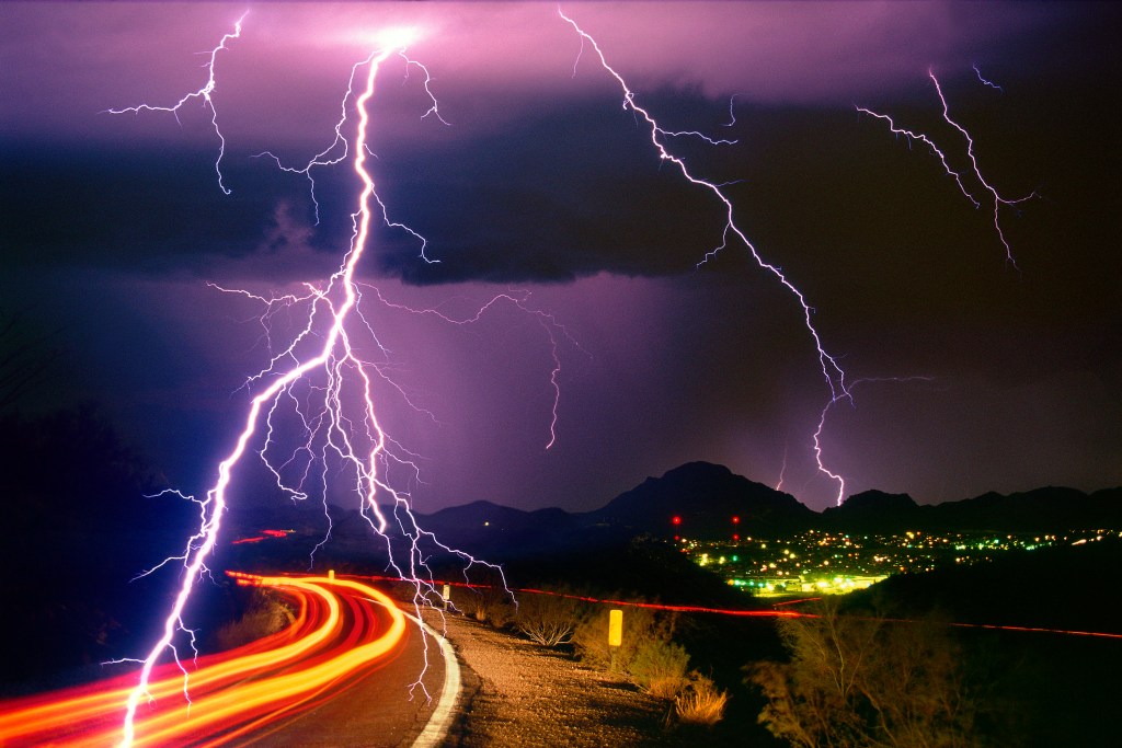 Sonoran Desert rainstorm, a summer monsoon thunderstorm with cloud-to-ground lightning strikes and light trails from cars on the roadway.