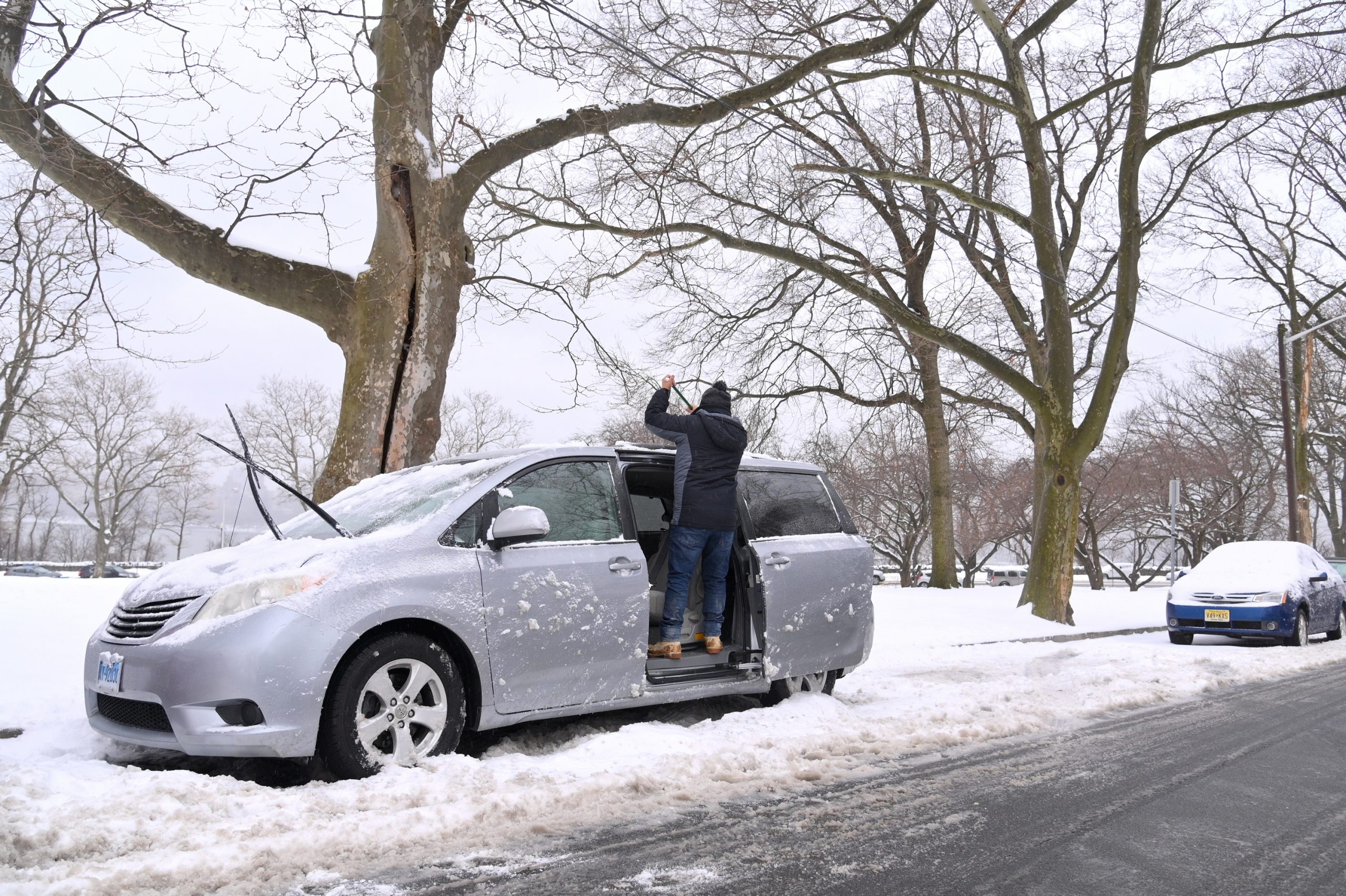A man cleans the snow off of his minivan parked on a street corner
