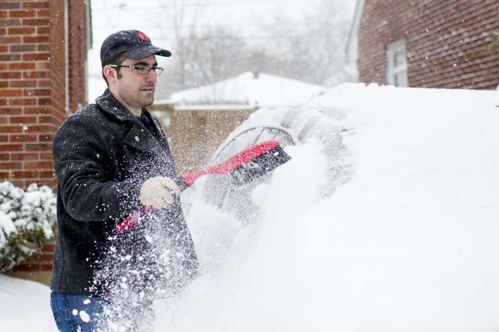 A man cleans snow from his car, Consumer Reports doesn't recommend warming up your car ahead of time.