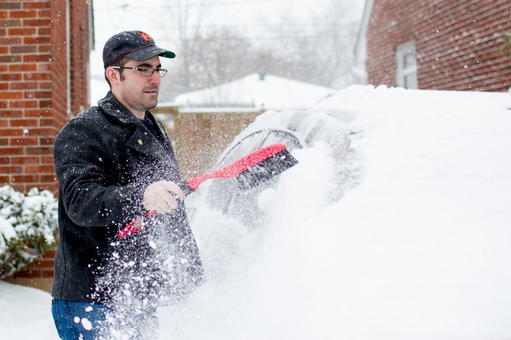 A man cleans snow from his car, a snow scraper is a good tip for driving in winter weather.