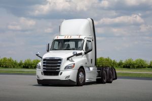 A white Freightliner Cascadia on a concrete drive with greenery behind it. 