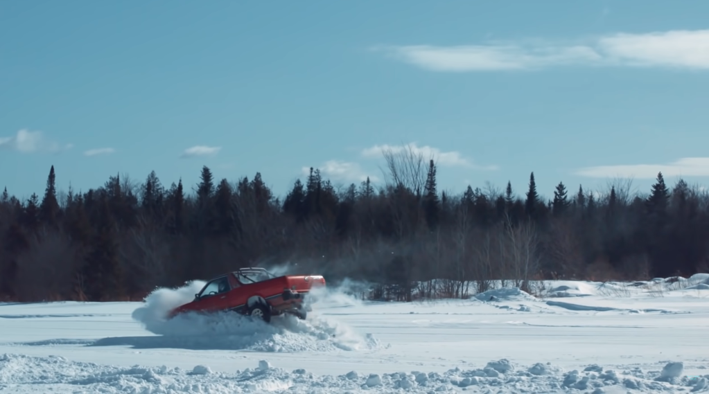A red 1984 Subaru Brat coupe utility lands in the snow after making a big stunt jump with Travis Pastrana behind the wheel.