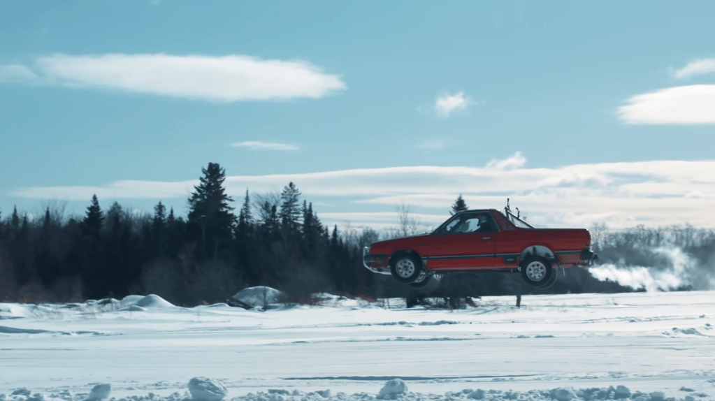 A red 1984 Subaru Brat coupe utility jumps over a frozen lake in Vermont with Travis Pastrana behind the wheel.