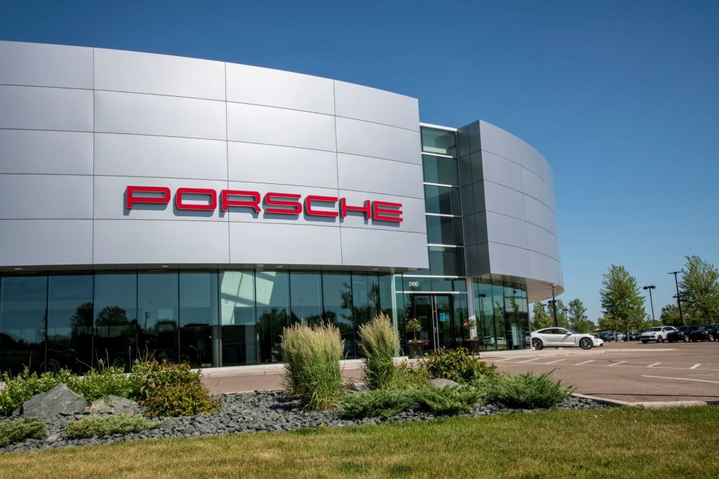 White Porsche dealership with Porsche written in red on the outside with grass and shrubbery.