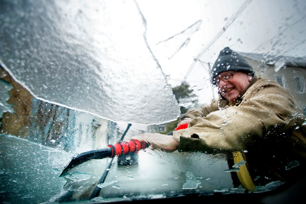 A man scrapes ice off the windshield of a car, an ice scraper is just one of our tips for driving in winter weather.