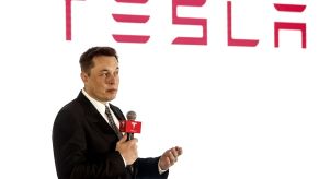 Tesla, creator of autopilot, speaking in front of a white background with Tesla written in red at the top dressed in a suit.