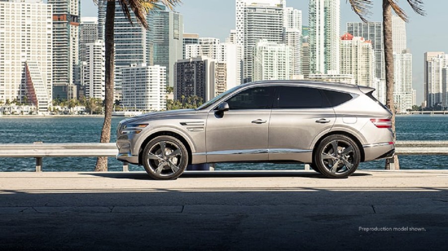 A silver Genesis GV80 driving with a city in the background.
