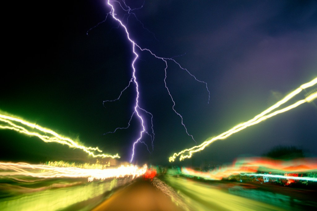 Nighttime storm on an interstate highway, with a lightning strike and light trails from passing cars and trucks. 
