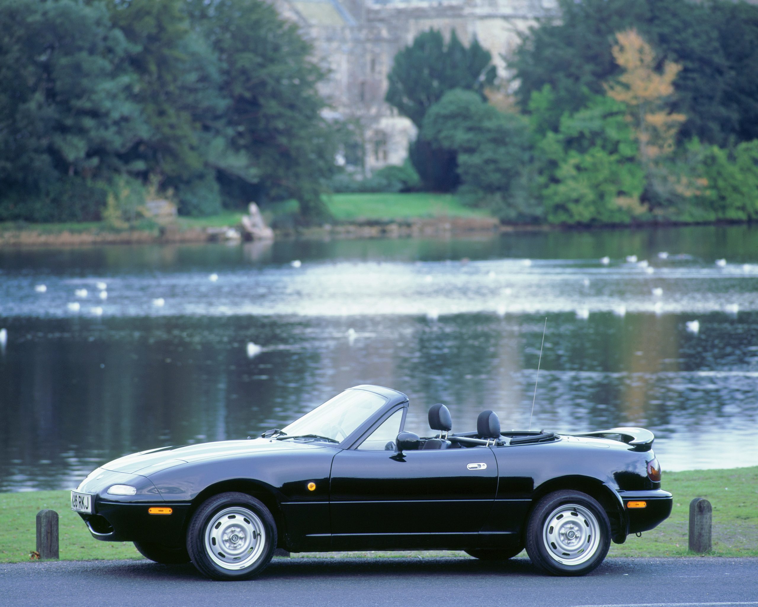A black NA generation Miata shot in profile in front of a lake in the UK