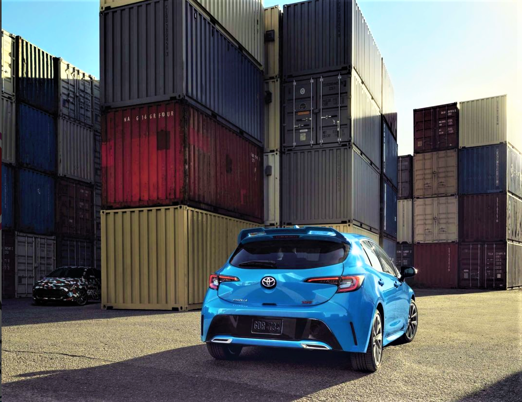 A rear 3/4 shot of a Toyota Corolla hides the new Toyota GR Corolla hot hatch in a camo livery among shipping containers