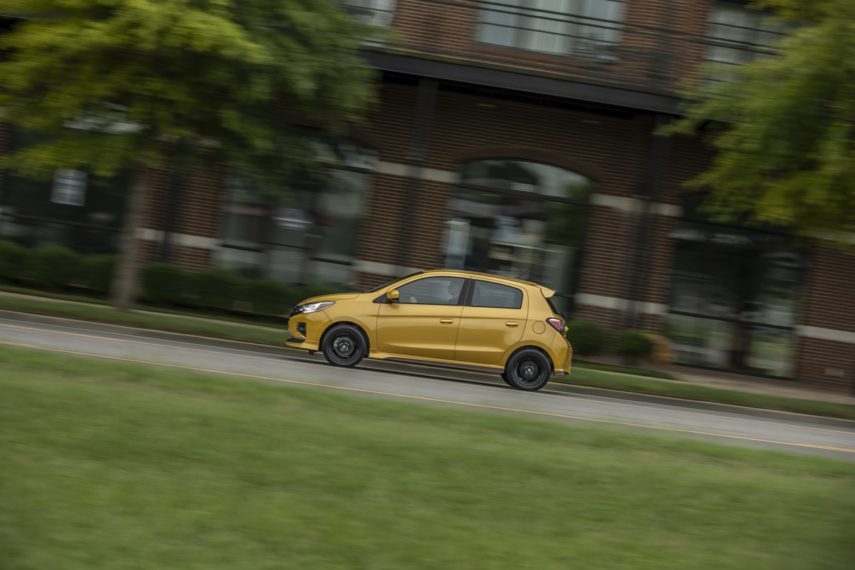 A yellow 2021 Mitsubishi Mirage subcompact hatchback traveling past a red-brick building, trees, and grass