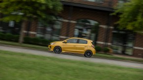A yellow 2021 Mitsubishi Mirage subcompact hatchback traveling past a red-brick building, trees, and grass