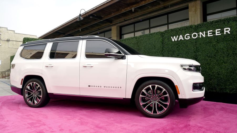 A white white Jeep Grand Wagoneer, maker of the Jeep Wagoneer trim, on a light red carpet in front of a black and brown building with Wagoneer printed in white on the side the building.