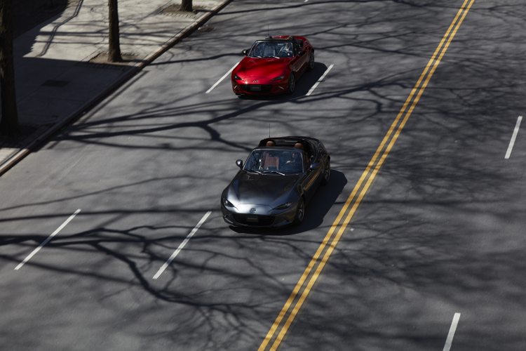 An overhead view of a gray Mazda Miata RF and a red Mazda MX-5 Miata convertible driving on a street.