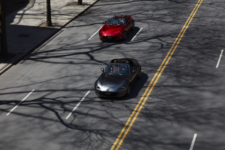 An overhead view of a gray Mazda Miata RF and a red Mazda MX-5 Miata convertible driving on a street.