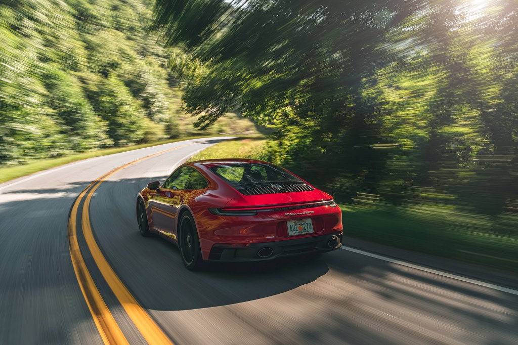 The rear 3/4 view of a 'Carmine Red' 2022 Porsche 911 Carrera GTS driving down a forest road