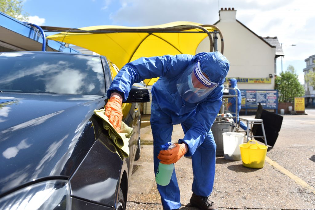 A person washing a car as part of a black car wash dressed in a blue hazmat style suit in an outdoor setting with a yellow tent in the background. 