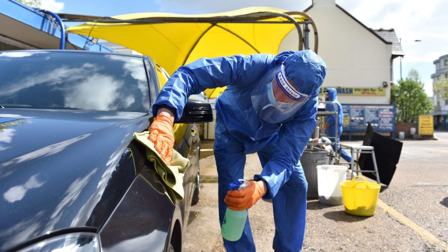 A person washing a car as part of a black car wash dressed in a blue hazmat style suit in an outdoor setting with a yellow tent in the background.