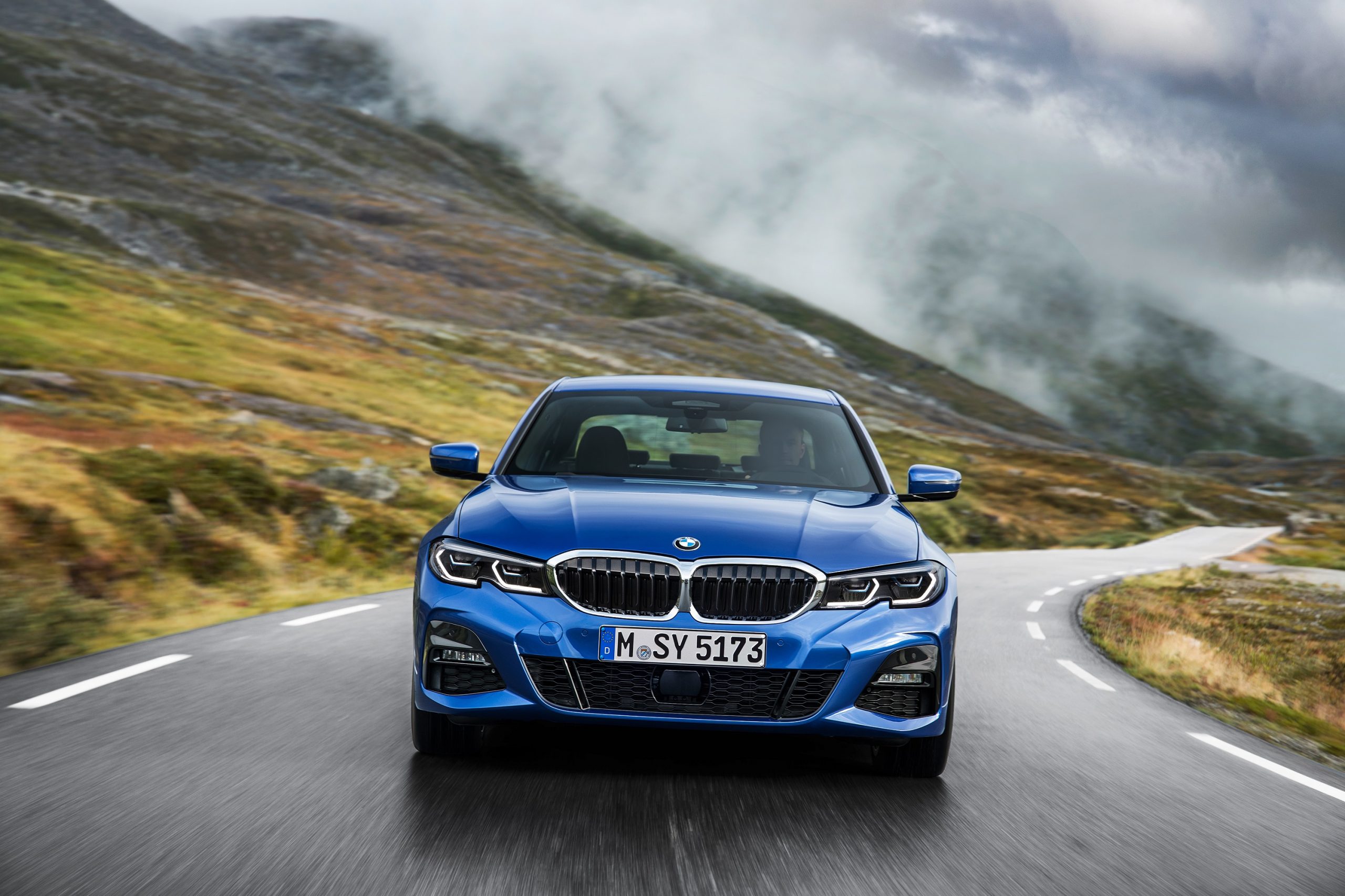 A blue 2019 BMW 3 Series sedan shot from the front on a Scottish B road