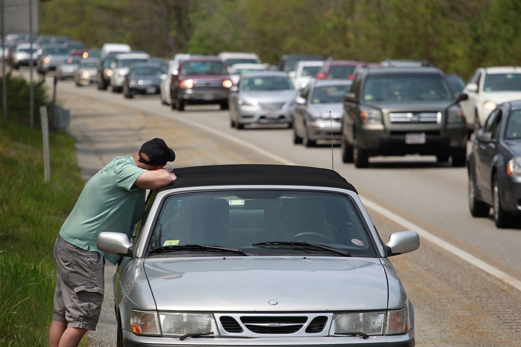A silver with a lack top breaks down on a highway with a person leaning against the car. 