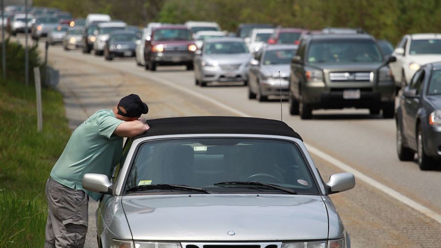 A silver with a lack top breaks down on a highway with a person leaning against the car.