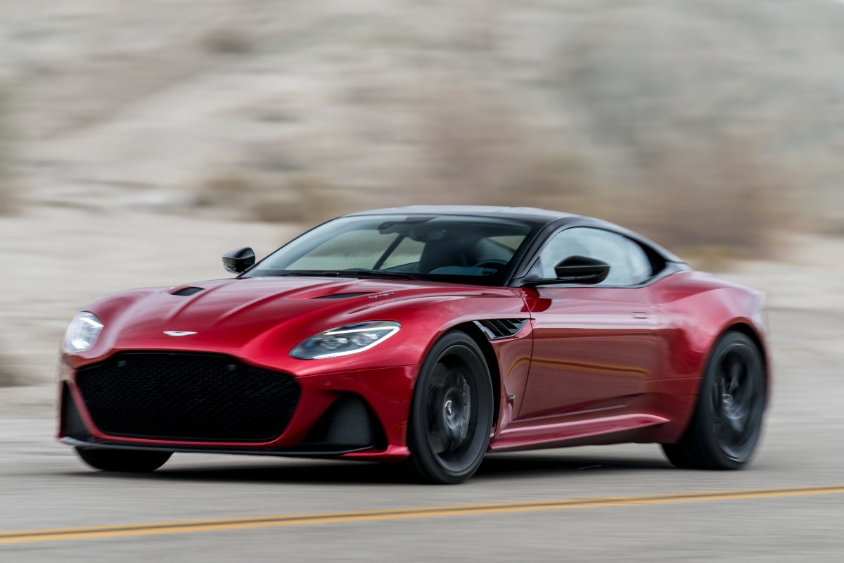 A red Aston Martin DBS driving fast on a road with rock cliffs in the background
