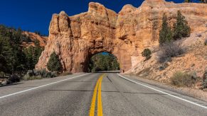 A road with a double solid yellow line in a rocky area with a tunnel ahead made of rocks.