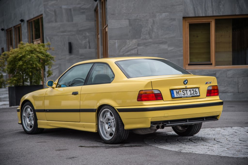 The rear 3/4 view of a yellow E36 BMW M3 Coupe next to a stone building