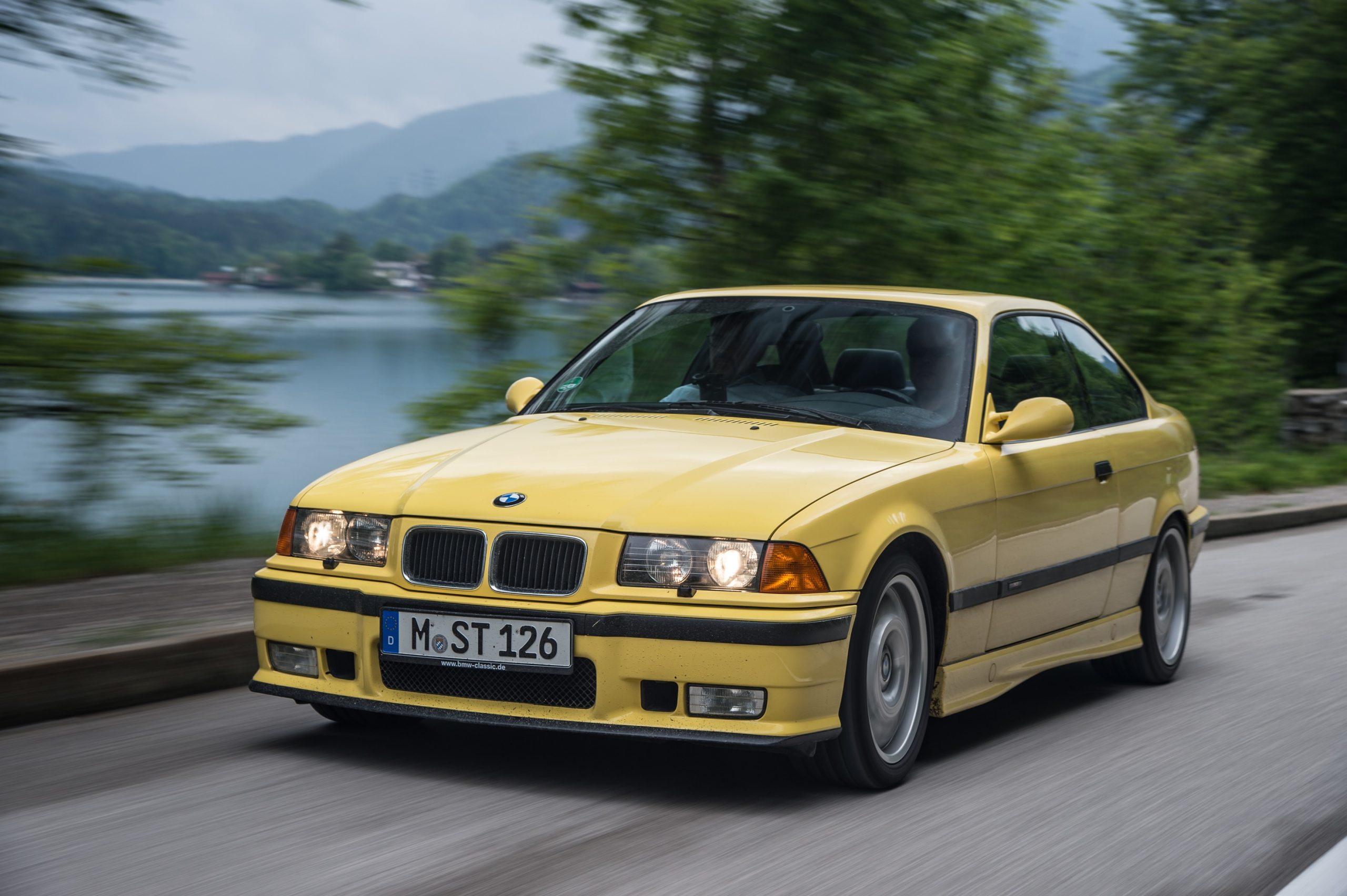 A yellow E36 BMW M3 Coupe drives around a mountain lake
