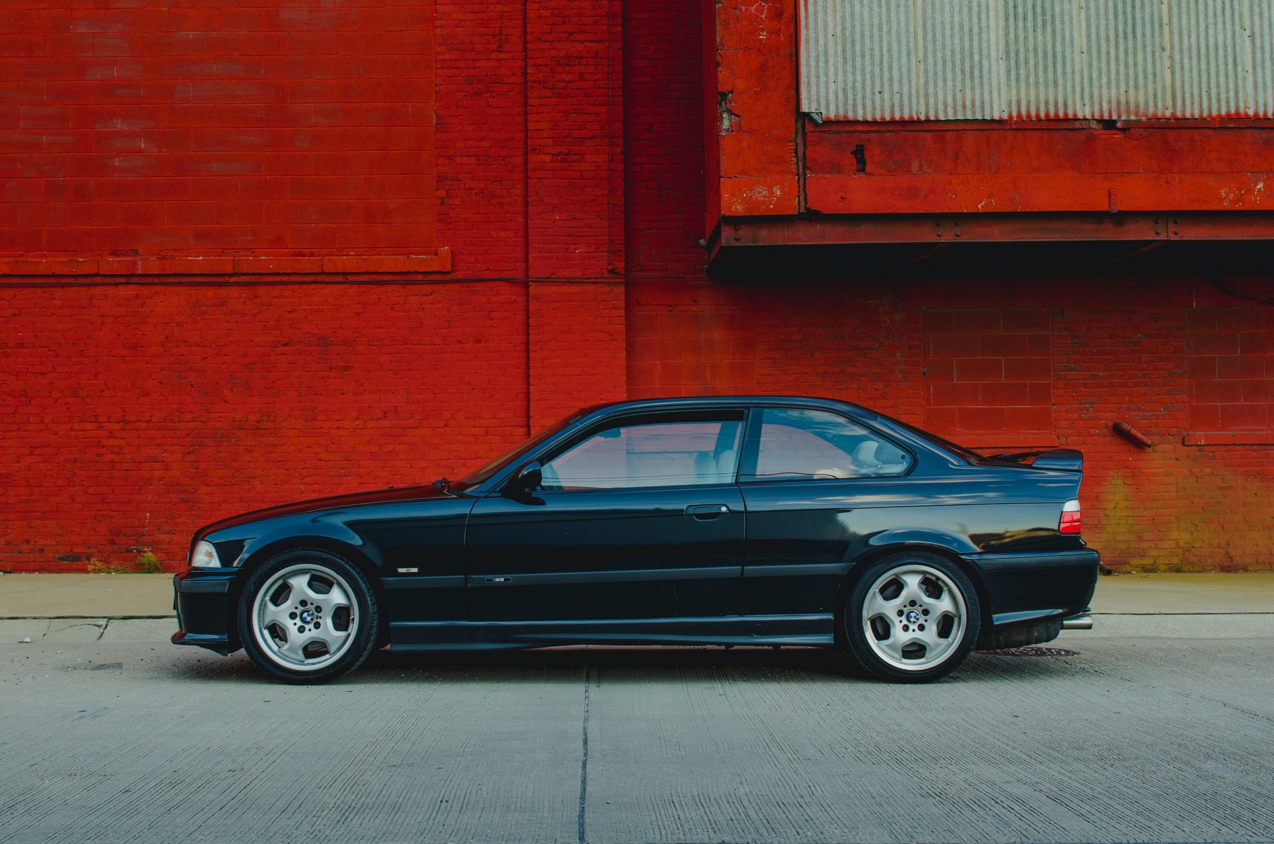 An E36 BMW M3 coupe shot in profile against a red brick wall