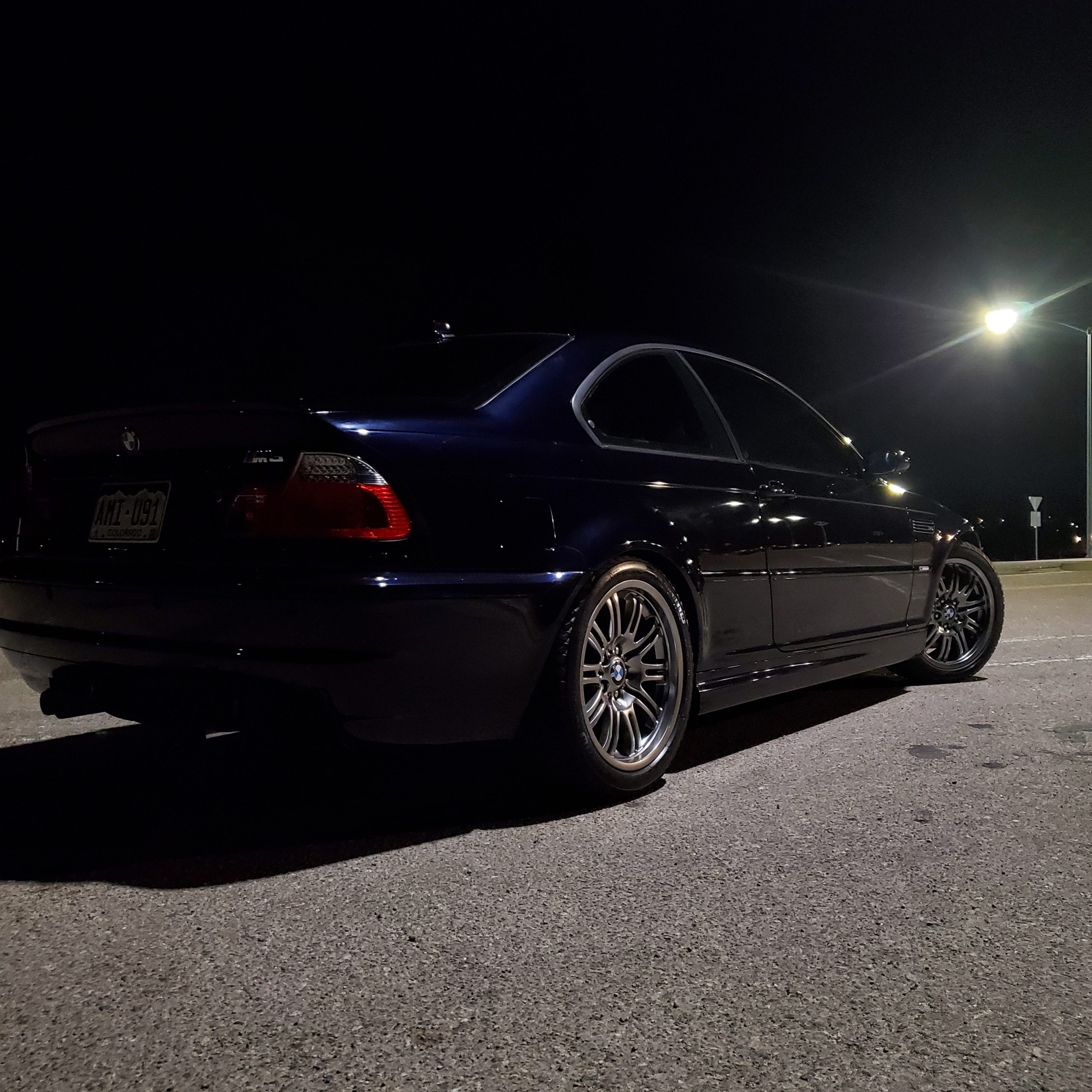 A black E46 BMW M3 shot from the rear 3/4 under a street lamp at night