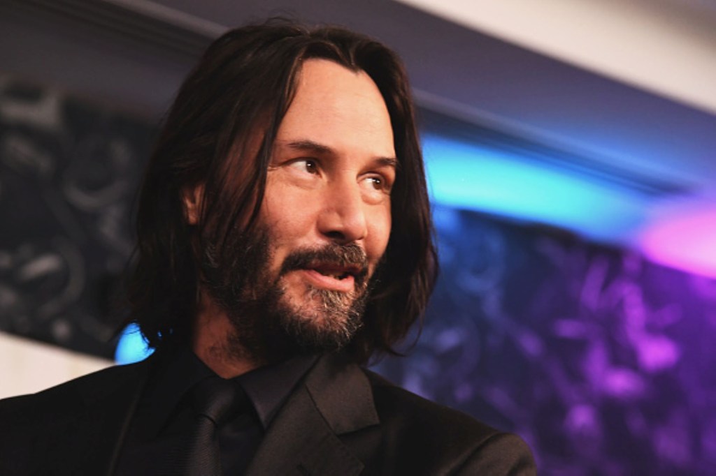 Keanu Reeves from 'John Wick' wearing a black suit with a black shirt and tie in front of a galaxy colored background with blues, purples, and blacks. 