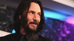 Keanu Reeves from 'John Wick' wearing a black suit with a black shirt and tie in front of a galaxy colored background with blues, purples, and blacks.