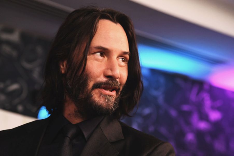Keanu Reeves from 'John Wick' wearing a black suit with a black shirt and tie in front of a galaxy colored background with blues, purples, and blacks.