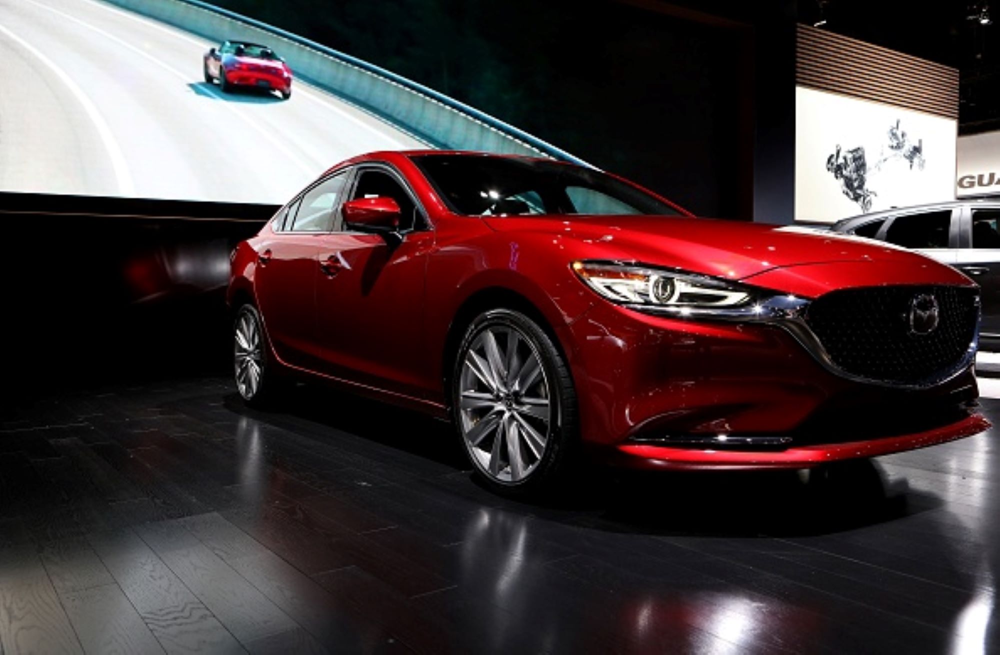 A red Mazda 6 parked in an indoor area with a screen in the background showing a highway with another Mazda 6 driving on it.