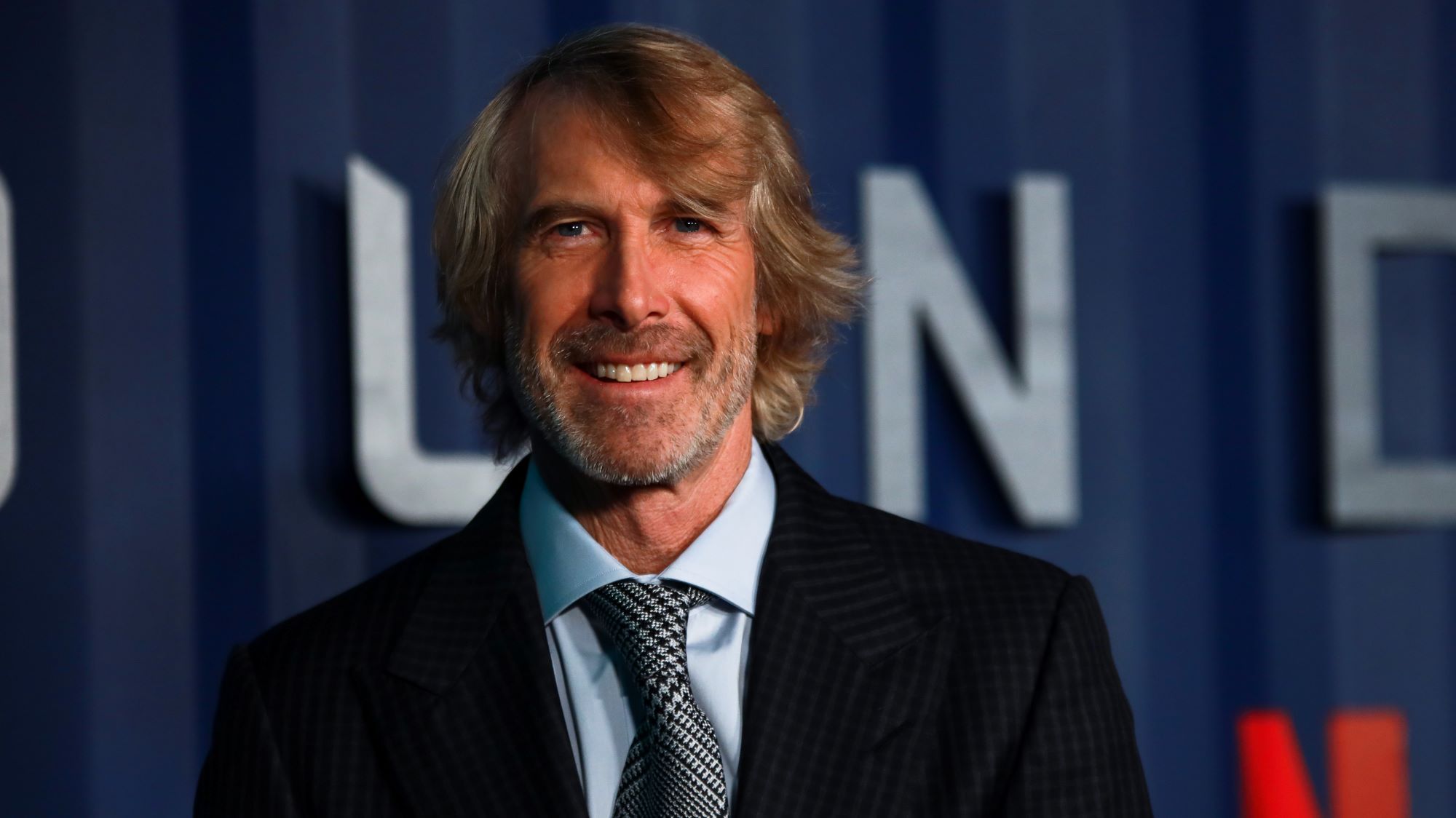 Michael Bay from 'Bad Boys' standing in front of a blue background with white letters dressed in a suit.