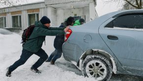 A group of people pushing a light blue car stuck in the snow.