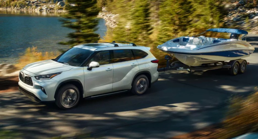 A white 2022 Toyota Highlander is driving on the road towing a small speed boat. 