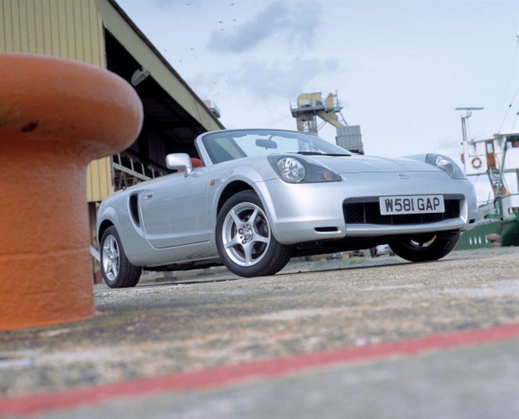 A silver Toyota MR2 in an outdoor environment with buildings around.