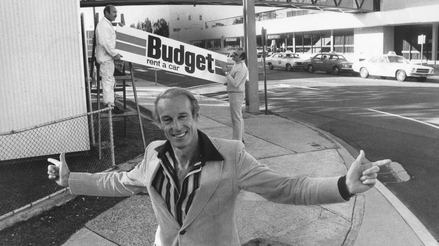 man standing in front of a sign reading "Budget car rentals" although the cost of a rental car has skyrocketed over the last few years.