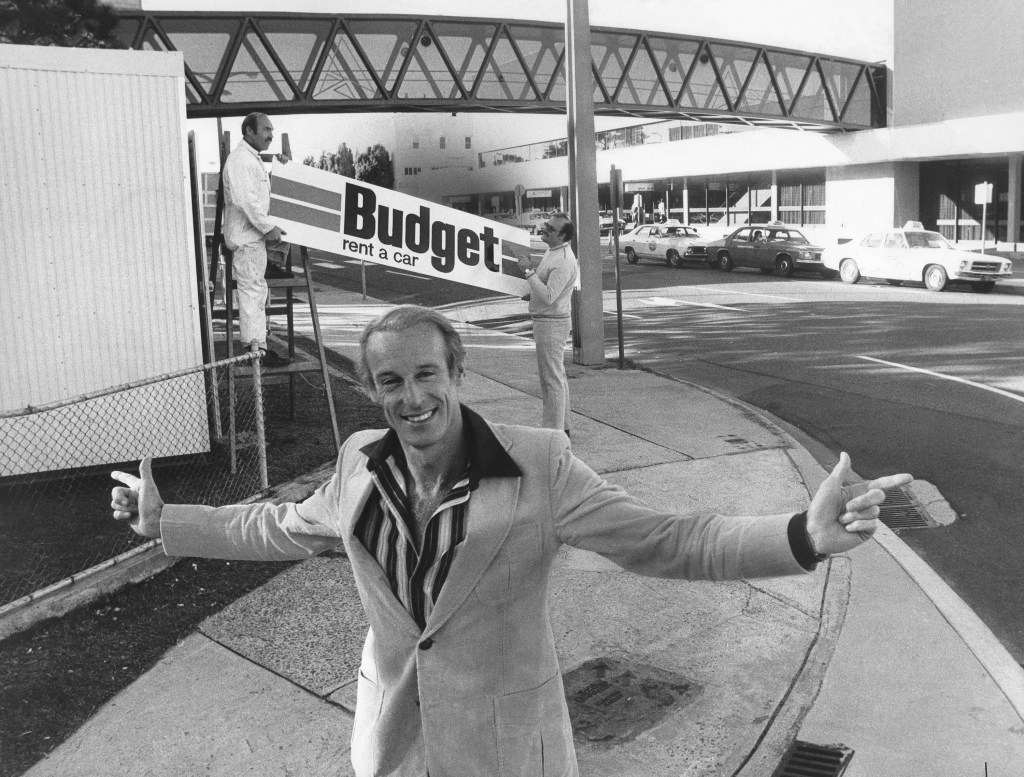 man standing in front of a sign reading "Budget car rentals" although the cost of a rental car has skyrocketed over the last few years. 