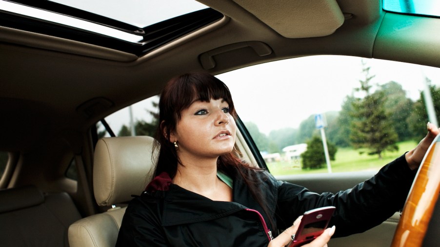 A young woman sits at the wheel of a car while holding a cell phone